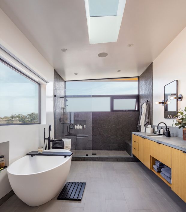 Contemporary spa-like bathroom with a freestanding white bathtub, large walk-in shower enclosed by glazing, double vanity with warm timber cabinetry and a dark benchtop, grey tiled walls and floor, skylight, and a large window overlooking greenery. Black fixtures and framed mirrors provide contrast.