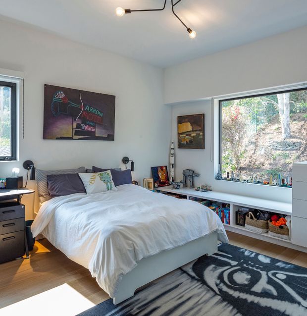Bright contemporary bedroom with a low upholstered bed dressed in white bedding, built-in shelving beneath a large window, bedside tables, artwork, and a black-and-white patterned rug. Natural light enters through the expansive window, with a leafy landscaped area visible outside. The room has pale walls, timber flooring, and a minimalist black branching ceiling light.