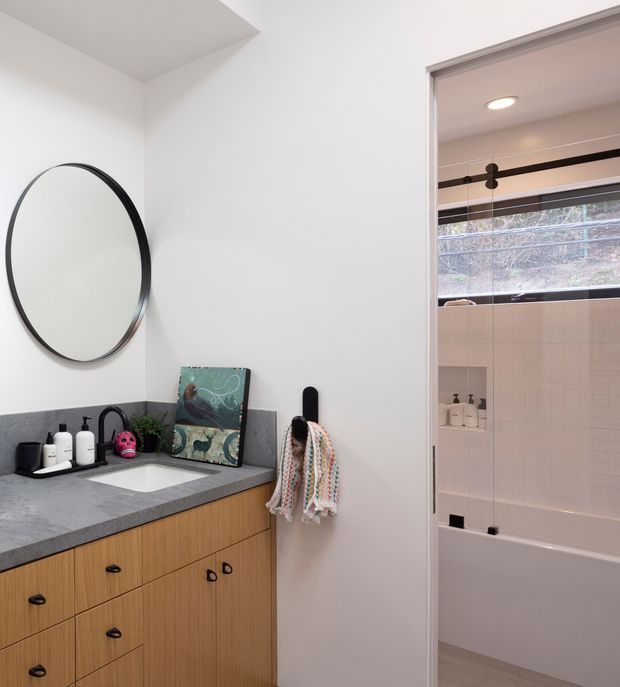 A bright, contemporary bathroom vanity area viewed from the room, with a round black-framed mirror, grey stone benchtop, white inset basin, and warm timber cabinetry. An open doorway reveals a bathtub with pale wall tiles, a horizontal window, and a dark exposed shower rail. Minimal accessories and white walls create a clean, calm atmosphere.