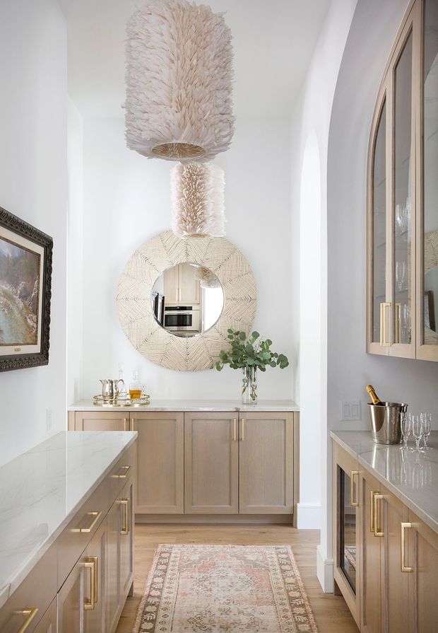 A bright, refined kitchen or butler's pantry with pale natural-wood cabinetry, light stone countertops, brass hardware, white walls, and warm timber flooring. A central island occupies the foreground, with a counter and cabinetry at the rear and right. Decorative pendant lights, a round woven-framed mirror, artwork, glassware, and a runner add a softly luxurious, layered feel. The mirror reflects additional kitchen appliances and joinery.