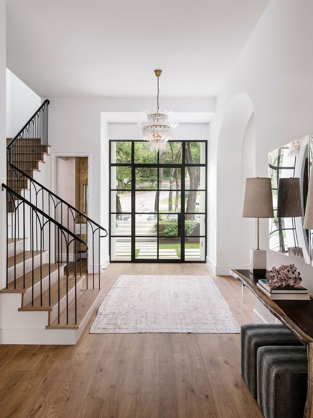 Bright, refined residential entry hall with a prominent timber staircase and slender black metal balustrade on the left. The space features white walls and ceiling, pale timber flooring, a large black-framed multi-pane glazed door opening to the outdoors, a crystal chandelier, a pale patterned rug, and a dark console with lamps and stools along the right wall. An arched opening adds architectural character.