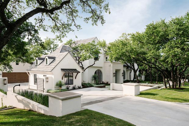 Exterior view of a refined white stucco residence with multiple pitched, shingle-clad roof forms, dormer windows, arched openings, and dark-framed windows. A broad concrete driveway and low masonry boundary walls lead through a mature, landscaped suburban setting with large established trees and manicured lawns.