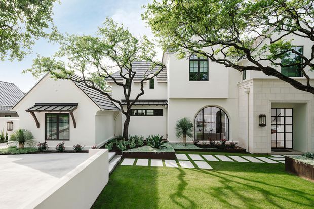 A bright landscaped courtyard frontage of a multi-wing residential home with white rendered walls, dark standing-seam pitched roofs, black-framed glazing, arched windows and doorways, and a formal lawn with large pale rectangular stepping slabs. Mature branching trees and sculptural drought-tolerant planting frame the facade.