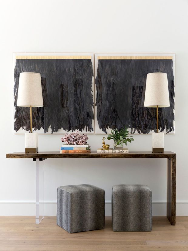 A refined interior vignette featuring a slim dark wood console table with clear acrylic supports, two grey upholstered cube stools, paired brass-and-cream table lamps, and layered black textured wall artworks. Decorative books, a small sculpture, and greenery style the console against a light wall and timber floor.