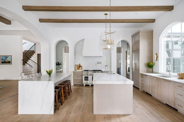 Bright, spacious kitchen with two large marble-look islands, pale wood cabinetry, arched openings, exposed timber ceiling beams, a decorative rangehood, and a large multi-pane window. The kitchen connects visually to a dining area and stairway, with a refined warm-neutral aesthetic.