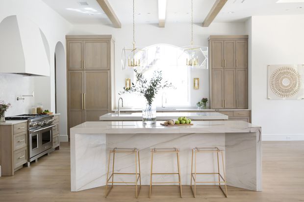 Bright, spacious kitchen with a large waterfall-edge island, three slim brass-framed stools, integrated sink, marble-look surfaces, tall timber cabinetry, stainless-steel range, arched openings, pendant lighting, exposed ceiling beams, and pale timber flooring. The room has a refined, warm contemporary aesthetic with abundant natural light.