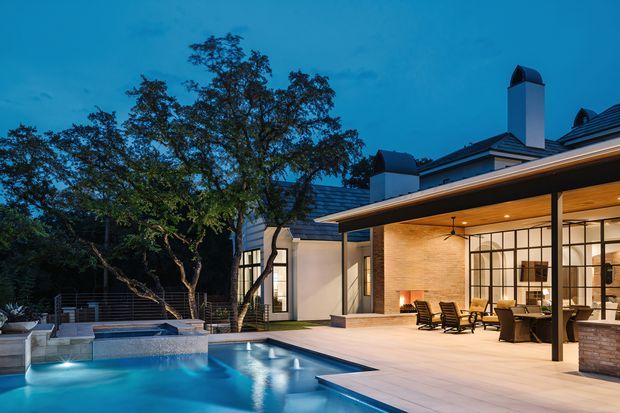 Twilight view of a refined contemporary home centred around a swimming pool and covered outdoor entertaining area. The architecture features pale rendered walls, dark pitched roof sections, prominent chimney forms, extensive black-framed glazing, and a broad timber-lined ceiling over the terrace. Outdoor seating and a fireplace create an inviting connection between the interior and pool deck.