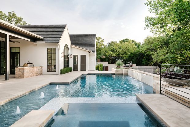 A contemporary white-rendered house opens onto a large landscaped swimming pool and shallow foreground water feature. The exterior has dark pitched tiled roofs, tall black-framed glazing, a covered outdoor kitchen or bar with a stone-clad island, pale paved terraces, raised planter beds, and landscaped greenery. The pool terrace is spacious, refined, and resort-like.