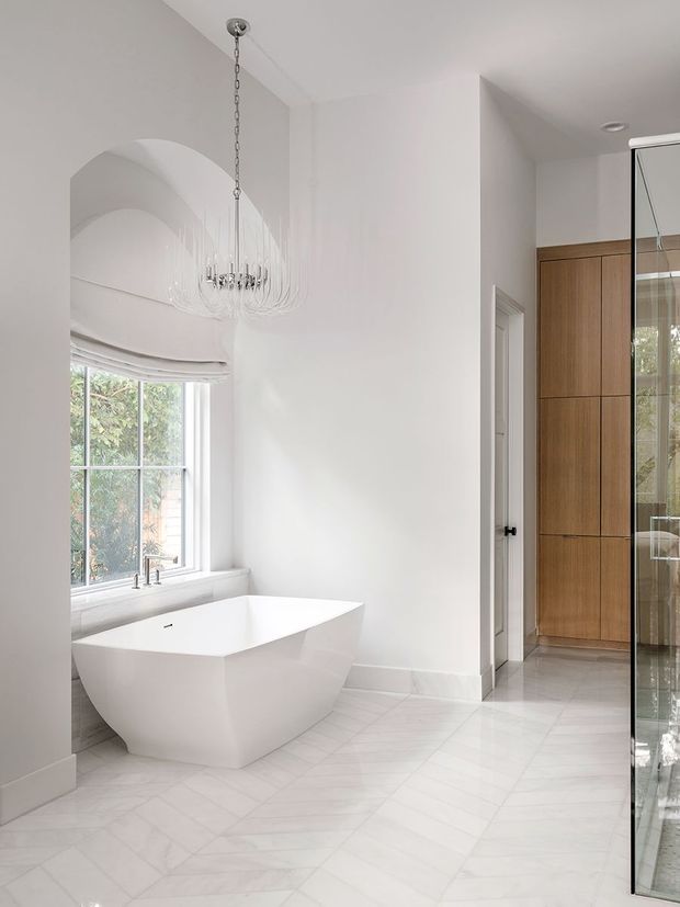 Bright, refined bathroom featuring a freestanding angular white bathtub beneath a large window, a sculptural hanging glass chandelier, white walls, and pale geometric stone or porcelain floor tiles. A timber-toned built-in vanity or storage wall and glass shower enclosure are visible to the right. The room has high ceilings and a clean contemporary luxury aesthetic.