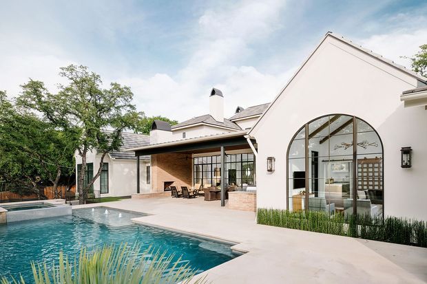 A bright courtyard-style residential exterior centered on a swimming pool and pale rendered buildings. The home has steep gabled roof forms, large black-framed arched and gridded glazing, covered outdoor living areas, brick detailing, and a landscaped pool terrace.
