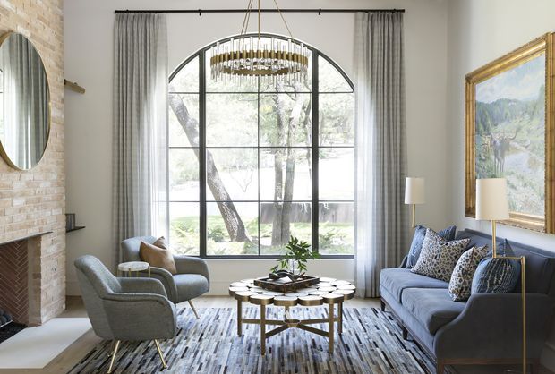 Elegant living room with a large arched, black-framed multi-pane window, full-height grey curtains, a blue-grey sofa and lounge chairs, layered coffee tables, patterned rug, brass chandelier, floor lamps, a brick fireplace wall, and framed artwork. The room has a refined, bright, traditional-contemporary character with strong natural light and garden views.