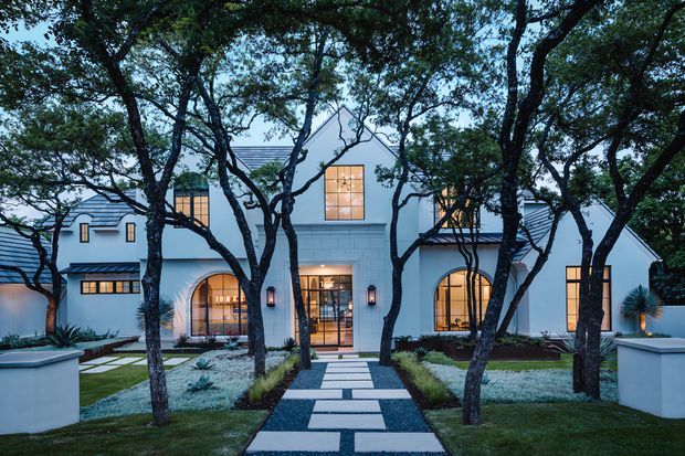 Front exterior view of a substantial white stucco residence framed by mature trees. The symmetrical facade features steep gabled roof forms, dark metal roofing, arched ground-floor windows, a central glazed entry, upper-level windows, and a formal landscaped garden with a geometric stepping-stone path.