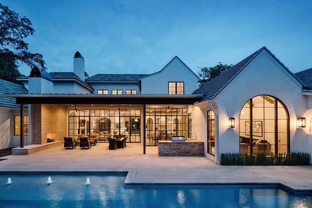 Twilight exterior view of a large single-storey residence surrounding a swimming pool and paved terrace. The home has white rendered walls, dark pitched and gabled roofs, prominent arched glazing, extensive black-framed windows, covered outdoor areas, and a brick-clad outdoor kitchen or bar counter. Warm interior lighting creates an inviting resort-like atmosphere.