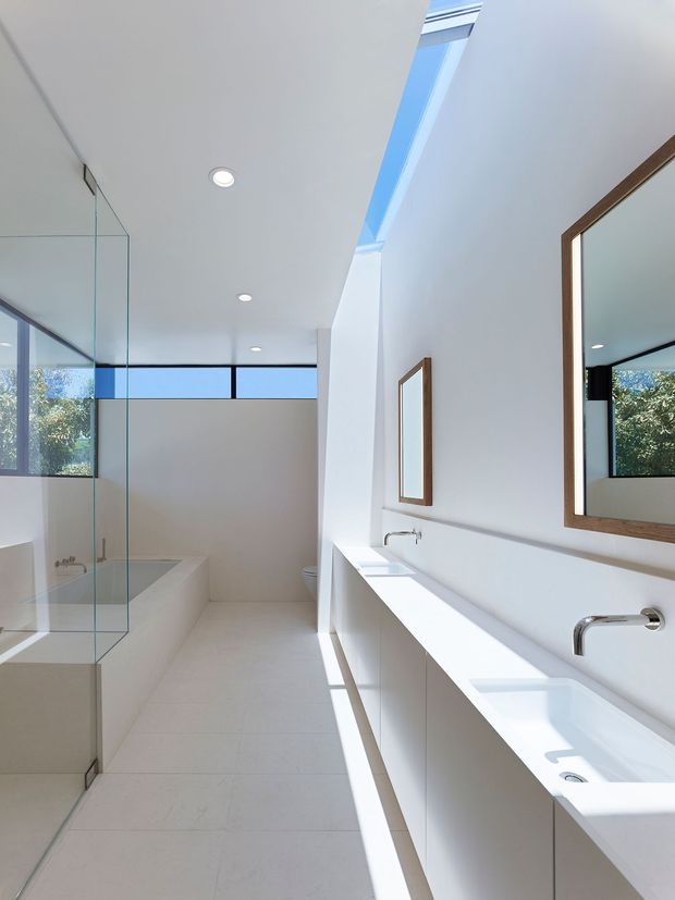 Bright contemporary bathroom with a long white double vanity, integrated basins, wall-mounted chrome faucets, large timber-framed mirrors, a glass-enclosed shower and freestanding bathtub. White tiled surfaces, recessed ceiling lights, clerestory glazing and a linear skylight create an airy, sunlit interior.