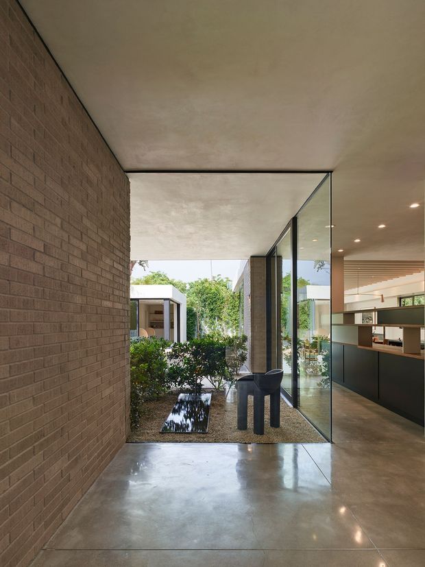 A contemporary interior corridor or entry space with a full-height exposed brick wall on the left, polished concrete flooring, and a large frameless glass wall opening onto a landscaped courtyard with a narrow reflecting pool. A dark sculptural chair sits beside the glazing. Through the glass, a sleek kitchen or living area is visible with dark built-in cabinetry, open shelving, and recessed lighting. The space has a restrained, material-focused indoor–outdoor design with strong visual connection to the garden.