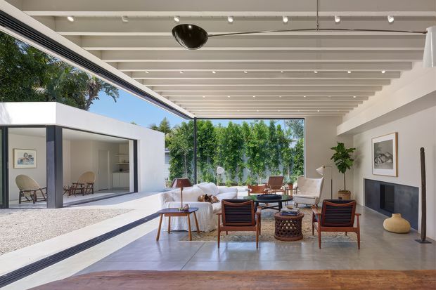 Bright contemporary living room opening onto a landscaped courtyard through extensive floor-to-ceiling glazing. The space features a white interior, exposed painted ceiling beams, polished concrete flooring, a fireplace with built-in ledge, and mid-century-inspired timber and upholstered furniture. A large framed opening connects visually to an adjacent pavilion or outdoor area.