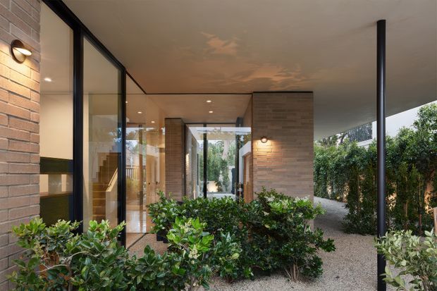 A contemporary covered residential entry or courtyard walkway with warm brick walls, slim black-framed floor-to-ceiling glazing, recessed and wall-mounted lighting, and a simple flat roof supported by slender black columns. Dense planted hedges line the gravel path, while a timber staircase and interior spaces are visible through the glazing.