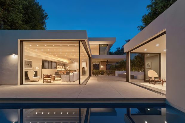 Twilight view of a low-slung contemporary residence arranged around a reflecting pool. Large glazed living spaces open onto a broad paved courtyard, with flat roof planes, slim dark frames, warm recessed and pinpoint ceiling lighting, and strong reflections in the pool. Trees form a subdued backdrop, while the interiors are visibly furnished with lounge seating and occasional chairs.
