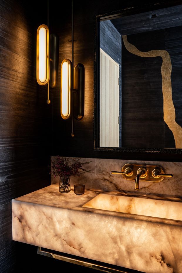 A dramatic, luxurious bathroom vanity with a floating stone basin and integrated backsplash. Dark textured wall cladding contrasts with warm translucent stone, brushed gold tapware, elongated wall sconces, and a large framed mirror reflecting a light-toned doorway or panel.