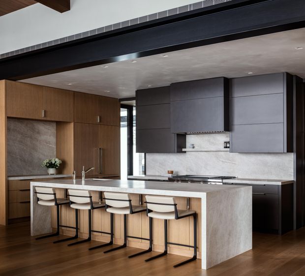 A sophisticated contemporary kitchen with a large stone island and four bar stools, dark grey built-in cabinetry, warm timber wall cabinetry, and a pale stone backsplash and benchtop. The room features timber flooring, recessed ceiling lighting, and a prominent exposed dark structural beam across the ceiling.