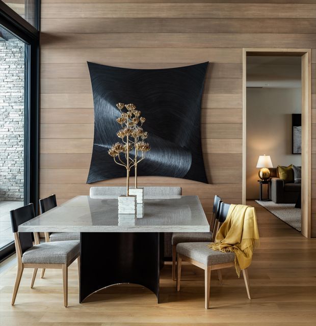 A refined dining area with a rectangular stone dining table, upholstered dining chairs, and a sculptural dark pedestal base. The room features horizontal timber wall panelling, timber flooring, black-framed glazing, and a large abstract black wall artwork. A doorway opens to a softly lit living room beyond, while a stacked-stone wall is visible through the glazing.