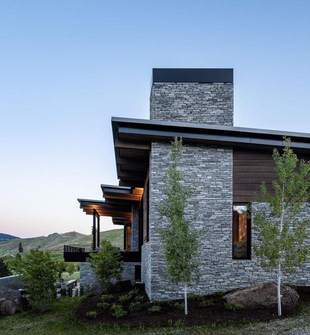 Exterior view of a contemporary multi-level residence set against a mountainous landscape. The building features a tall stacked-stone chimney or feature wall, dark horizontal timber cladding, broad projecting roof eaves, deep covered outdoor terraces, slender dark posts, and large windows. Landscaping with young trees, shrubs, and boulders surrounds the house.