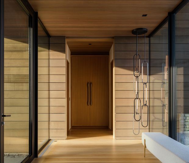 A warm, wood-lined entryway with full-height glazing and dark-framed sliding doors. Horizontal timber wall cladding and a timber ceiling frame a recessed built-in storage or entry cupboard with tall double handles. A sculptural black pendant hangs beside the entry, and the corner of a light upholstered bench is visible in the foreground. Strong natural sunlight creates a refined, calm atmosphere.