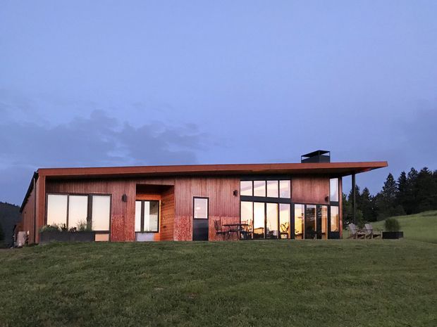 A low, single-storey contemporary timber-clad house set in an open grassy landscape at dusk. The long horizontal form has a shallow flat roof with a small rooftop flue or vent, extensive floor-to-ceiling glazing, and a recessed timber entry. Warm interior light is visible through the large windows, with a small outdoor dining setting on the covered terrace.