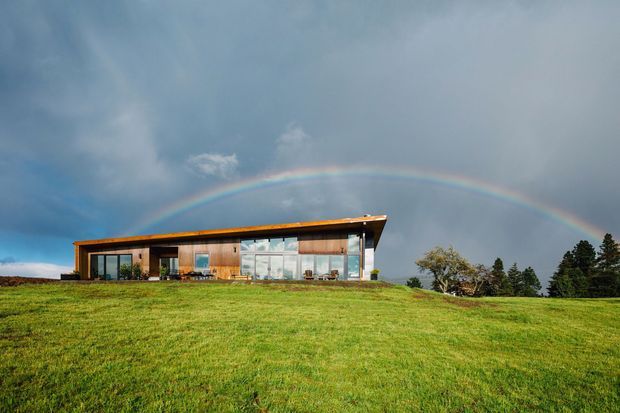 A low-slung contemporary single-storey house set on an open grassy hillside. The building has extensive floor-to-ceiling glazing, warm timber-clad sections, a long projecting roof with a shallow pitch, and a simple horizontal form. A rainbow arcs above the house, with scattered trees in the background.