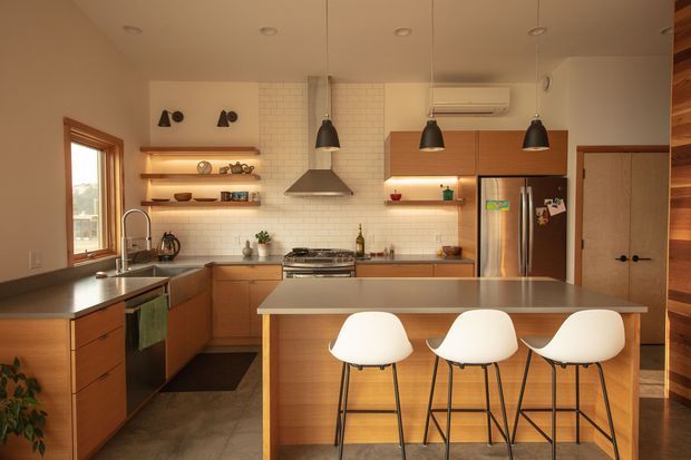 Warm, well-appointed contemporary kitchen with extensive timber cabinetry, a large central island with three stools, stainless-steel appliances, white tiled splashback, open display shelving, pendant lights, and an adjacent timber-lined passage or pantry area.