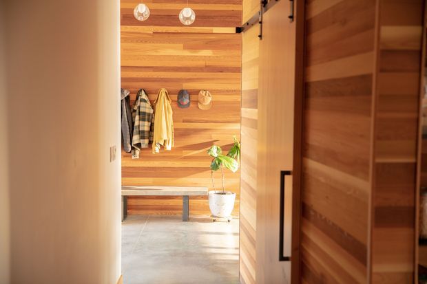 View through an open doorway into a warm timber-lined entry or mudroom. Horizontal wood wall and door cladding, exposed dark metal hardware, a simple bench with coat hooks, hanging jackets, hats, a potted plant, and a smooth grey concrete floor create a compact, practical space with strong natural-material character.