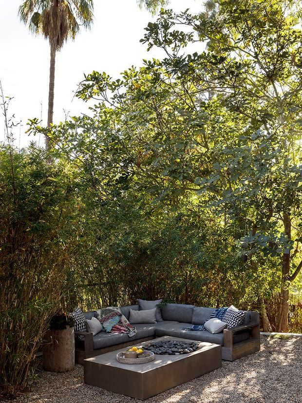 A landscaped outdoor lounge set within dense tropical greenery, featuring a dark grey sectional sofa with patterned cushions arranged around a low rectangular fire-pit table. The ground is covered in light gravel, with large planters and mature palms and foliage creating a private, garden-like setting. The scene is warmly lit and inviting.