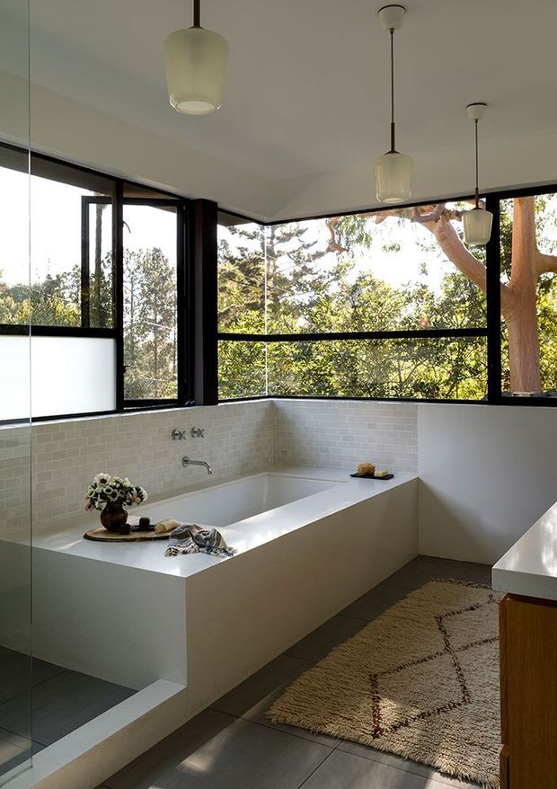Bright contemporary bathroom with a built-in white rectangular bathtub beneath expansive black-framed windows overlooking trees. White small-format tiled walls, dark grey floor tiles, a frameless glass shower panel, pendant lights, and a textured neutral rug create a clean, light-filled interior.