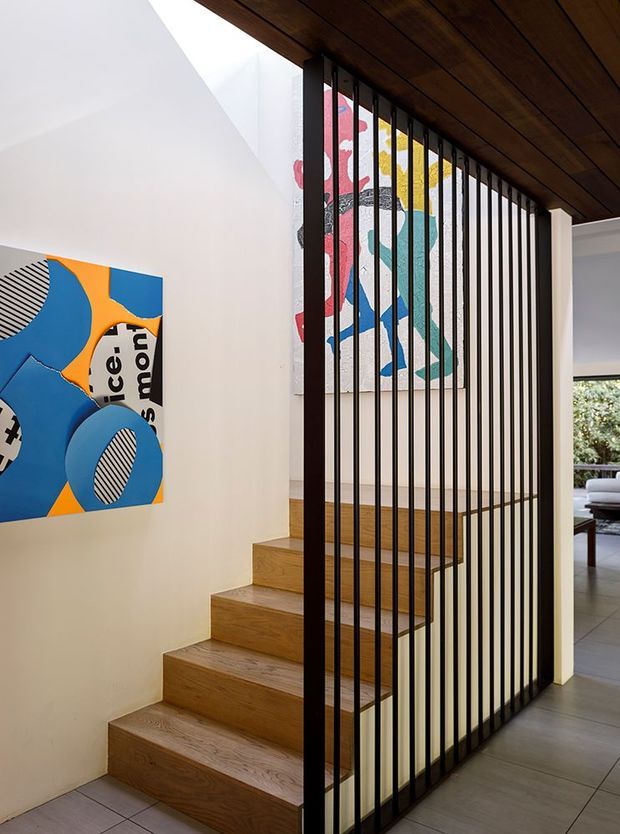 Interior view of a contemporary residential stairway featuring warm timber treads and a dark timber ceiling, enclosed by a tall black vertical slat screen and framed by white walls. Colourful abstract artwork adds visual interest, with a glimpse of an adjacent living area beyond the screen.