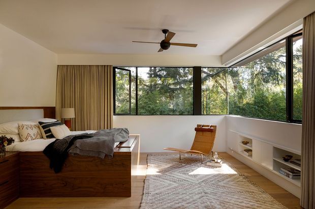 A warm, mid-century-inspired bedroom with a low built-in timber bed platform and integrated bedside storage, a neutral upholstered bed, wood-panelled curtains, expansive black-framed corner windows, a ceiling fan, built-in white shelving, and a woven area rug. The room overlooks leafy greenery and has a calm, light-filled atmosphere.