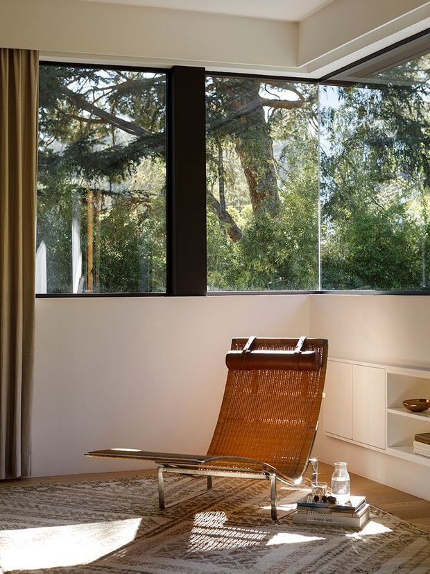 A refined sitting area with floor-to-ceiling corner glazing overlooking mature trees. A woven leather lounge chair sits on a patterned rug beside low white built-in shelving, with warm curtains, pale walls, timber flooring, and strong natural sunlight creating a calm, understated atmosphere.