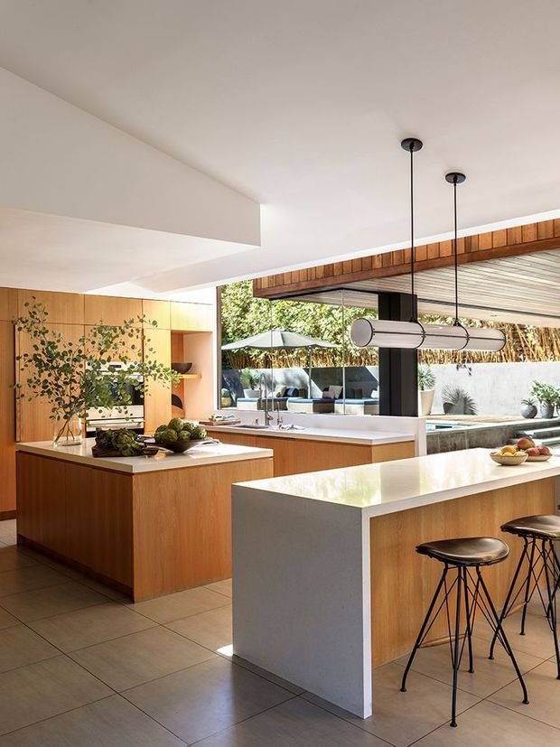 Bright contemporary open-plan kitchen with extensive warm timber cabinetry, two large white stone-look islands, integrated appliances, and black bar stools. The room opens directly to a landscaped outdoor area through large glazed openings, with timber ceiling detailing and linear pendant lights. Neutral tiled flooring and abundant natural light create an airy indoor-outdoor setting.