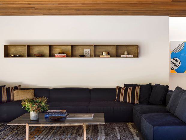 A contemporary living room with a large dark charcoal L-shaped sectional sofa, patterned cushions, a timber-and-metal coffee table, and a textured area rug. A long recessed timber display shelf runs along the white wall, while the ceiling is clad in warm timber boards. The room has a refined, warm, high-contrast aesthetic with a small glimpse of artwork at the right edge.