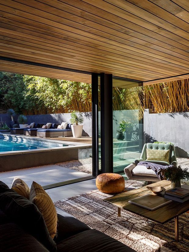 A warm, contemporary living room opens through large black-framed glazed doors onto a landscaped pool terrace. The room features a dark sofa, low timber coffee table, patterned rug, sculptural leather ottoman, and upholstered lounge chair. A timber-lined ceiling extends over the indoor-outdoor area, while a rendered privacy wall, bamboo planting, and swimming pool are visible outside.