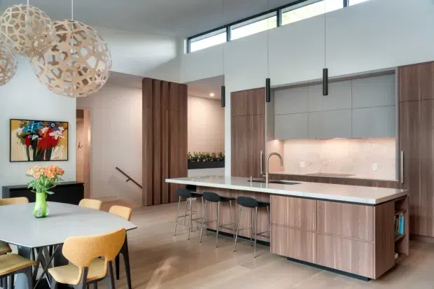 Open-plan contemporary kitchen and dining area with a large timber island, pale stone benchtop, integrated cabinetry, pendant lighting, and a high clerestory window. A dining table and chairs occupy the foreground, while a staircase and living area are visible beyond the kitchen.