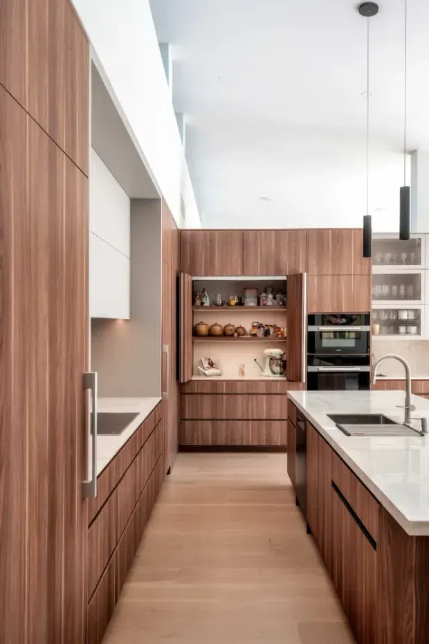 A bright, contemporary galley-style kitchen with extensive warm walnut-toned built-in cabinetry, integrated appliances, a central island with a white stone benchtop and undermount sink, open shelving, and slim black pendant lights. Light timber flooring and white walls create a clean, refined interior.