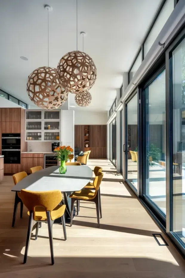 Bright, contemporary open-plan kitchen and dining area with extensive floor-to-ceiling glazing along one side. The space features warm timber cabinetry and flooring, a large dining table with mustard-yellow upholstered chairs, a built-in kitchen with appliances and display shelving, and sculptural woven pendant lights. Strong natural sunlight creates an airy, inviting atmosphere.