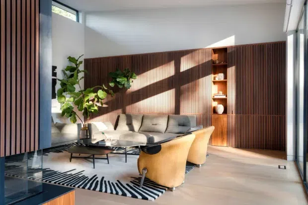A bright, contemporary living room with a grey sectional sofa, two mustard-yellow lounge chairs, a black coffee table, and a black-and-white striped rug. A full-height dark timber slatted feature wall incorporates built-in cabinetry and open shelving, while large windows and clerestory glazing bring in strong natural light. Indoor plants and carefully integrated joinery create a refined, warm interior.