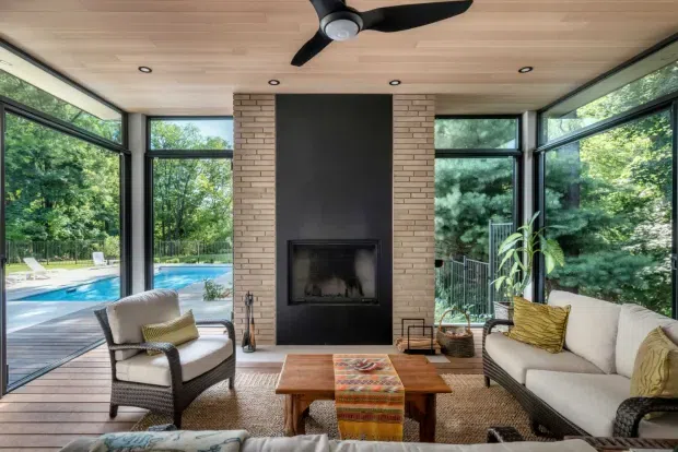 A bright, glass-enclosed living room overlooking a swimming pool and leafy landscape. The room features a central dark fireplace set between exposed light brick piers, pale timber ceiling and flooring, expansive black-framed glazing, a ceiling fan, a white sofa, woven armchair, timber coffee table, and neutral textured furnishings with colourful patterned accents.