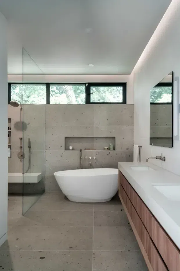 Contemporary bathroom featuring a freestanding white bathtub, large-format grey concrete-look wall and floor tiles, a frameless glass shower enclosure, a long white double vanity with warm timber cabinetry, recessed wall niches, black-framed clerestory windows, and a large wall mirror. The room is bright, clean, and minimalist with natural greenery visible through the windows.