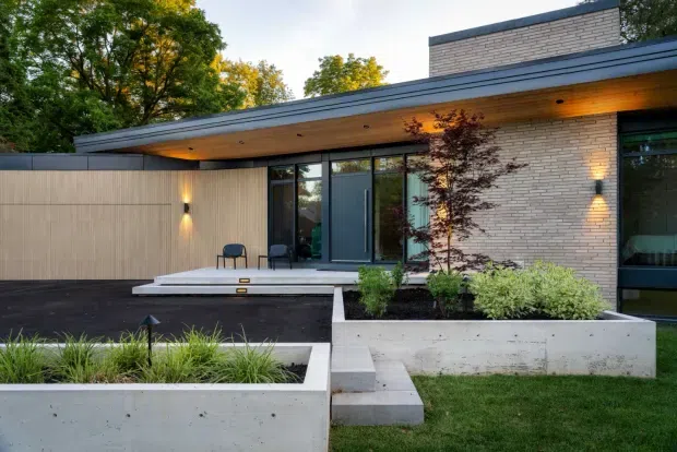 Exterior view of a low-slung contemporary residence with a broad flat roof and deep timber-lined eaves. The facade combines pale brick, dark-framed glazing, vertical timber screening, and exposed concrete raised planters and steps. A landscaped front garden with ornamental grasses and shrubs sits in the foreground, with warm integrated wall and soffit lighting creating an inviting evening atmosphere.