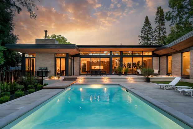 A low-slung, single-storey modern residence arranged around a rectangular swimming pool and paved terrace. The dark horizontal rooflines, extensive floor-to-ceiling glazing, warm timber soffits, and illuminated interiors create a refined evening setting. Mature trees frame the house, while broad steps lead from the living spaces down to the pool deck.