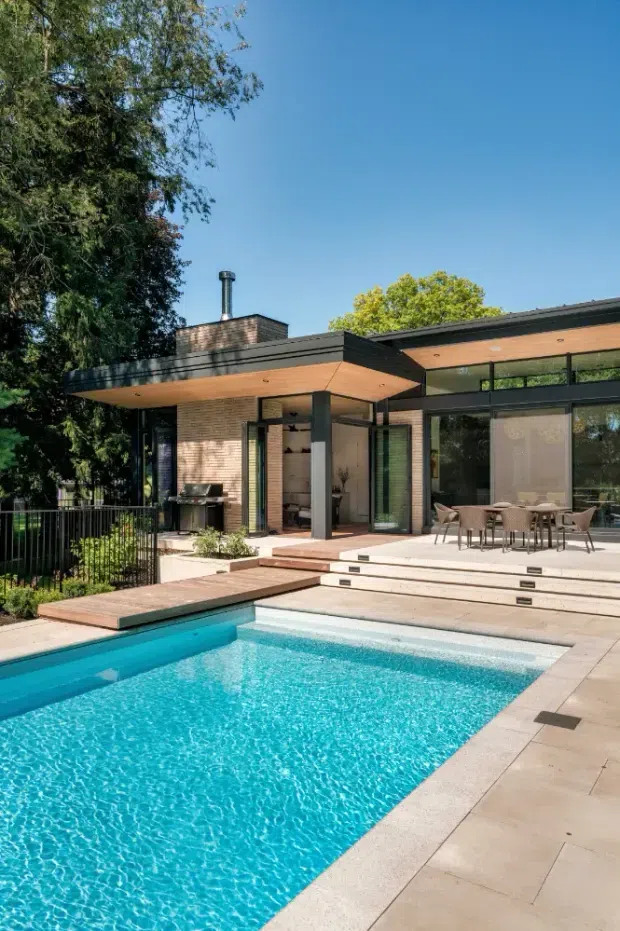 A modern single-storey house opens onto a stepped outdoor terrace and bright rectangular swimming pool. The facade combines dark horizontal roof and frame elements with warm timber soffits, pale brick walls, and extensive floor-to-ceiling glazing. A covered outdoor dining area, barbecue, deck, planted garden edges, and mature trees surround the pool.