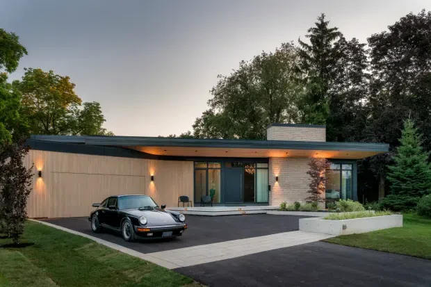 Single-storey modern residence viewed from the driveway at dusk, with a broad flat roof, warm vertical timber-clad facade, pale brick feature wall, dark-framed glazing, integrated exterior lighting, and a minimalist paved approach. Mature landscaping surrounds the home, and a classic car is parked in the forecourt.