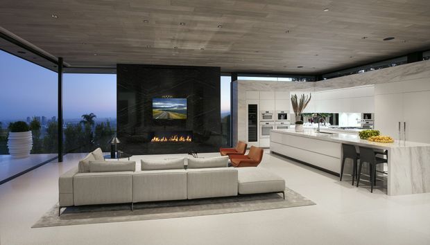 An expansive open-plan contemporary interior combining a living room and kitchen. A large pale sectional sofa faces a dramatic dark stone fireplace with a linear gas flame and television, while the bright white kitchen has a long island with integrated seating and extensive built-in cabinetry. Floor-to-ceiling glazing opens to a distant landscape, and the room features a continuous timber-lined ceiling, polished light flooring, and minimalist detailing.